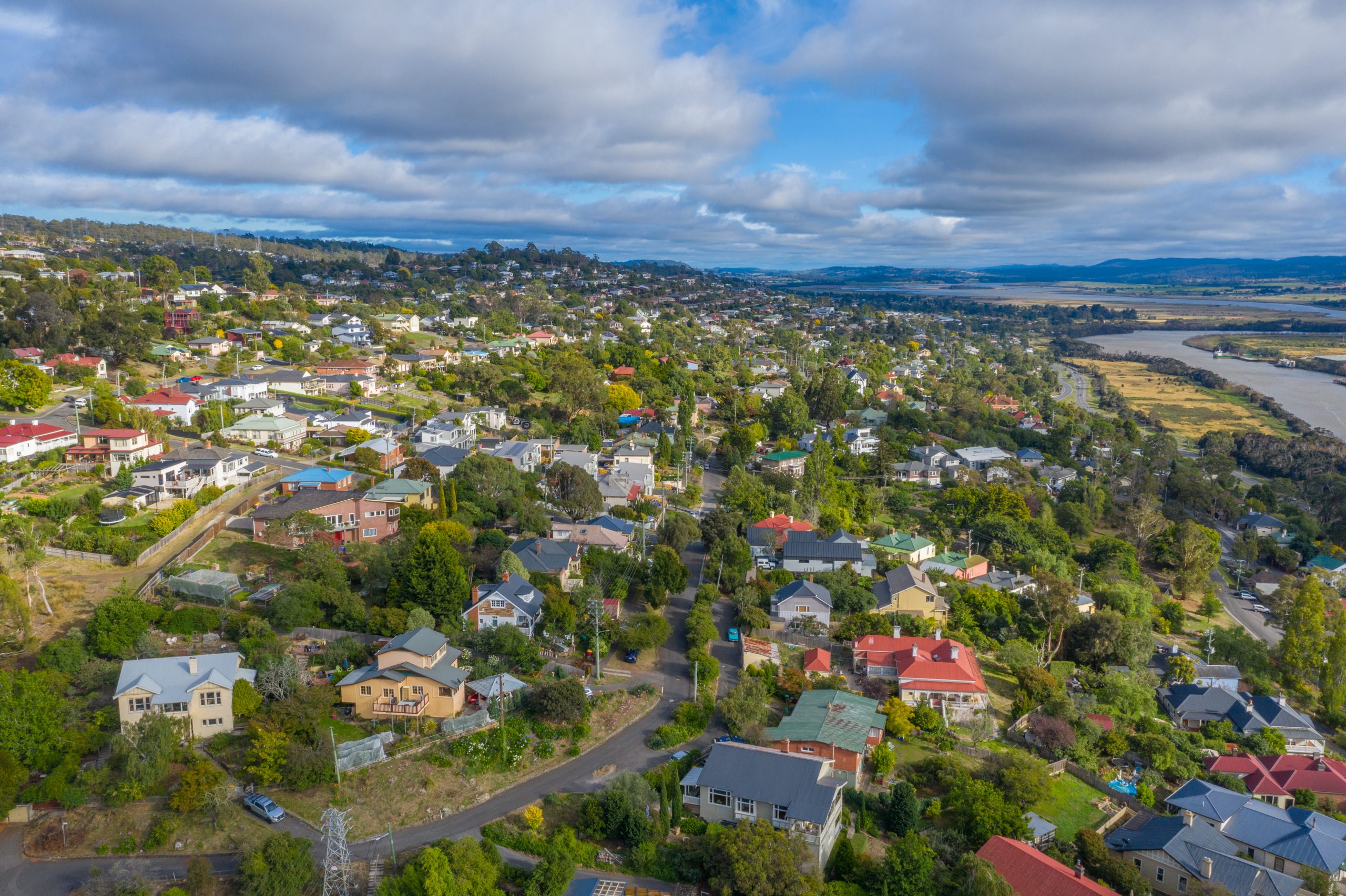 Aerial view of a tree-lined Tasmanian residential suburb with mature canopy cover, with the Tamar River and bushland in the distance.